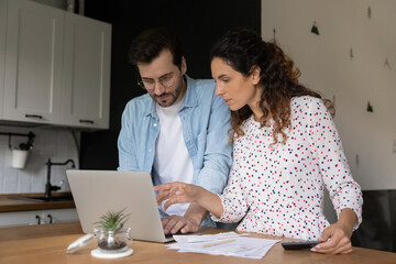 Young Caucasian couple stand at kitchen counter look at laptop screen pay bills taxes online. Millennial man and woman calculate household family expenses, make payment online on computer at home.