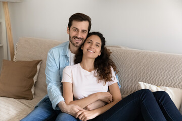 Portrait of happy young Caucasian man and woman sit rest on couch at home, show love and care in relationship. Smiling couple renters or tenants relax in new own house. Realty, moving concept.