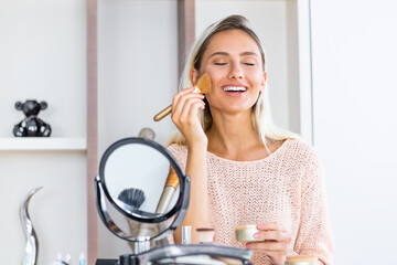 Beauty woman applying makeup. Beautiful girl looking in the mirror and applying cosmetic with a big brush. Girl gets blush on the cheekbones. Powder, rouge