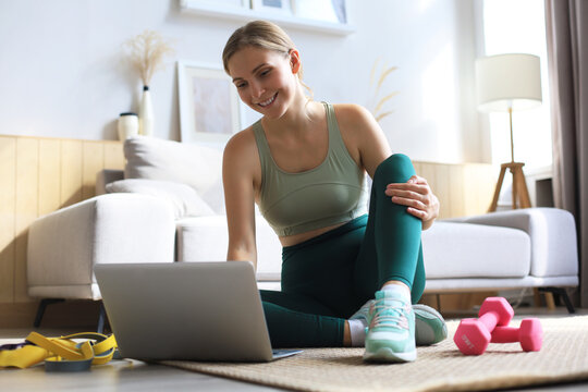 Beautiful slim sporty woman in sportswear is sitting on the floor with dumbbells and is using a laptop at home in the living room. Healthy lifestyle. Stay at home activities.