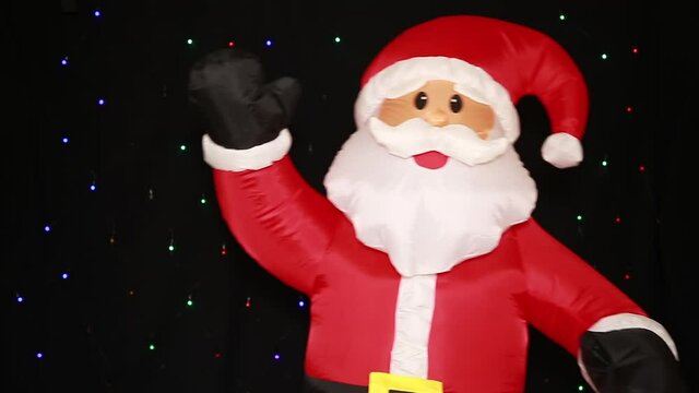 A large inflatable life-size Santa Claus doll, in a red suit, swaying merrily, waving a hand in greeting, isolated on a black background with flashing lights. Happy New year celebration happy holiday