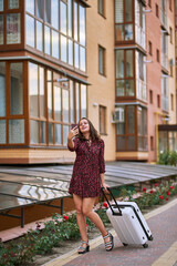 A girl in a red dress stands in the yard of the building with a white suitcase and is about to leave. she thinks walks around the suitcase