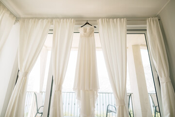 Fototapeta na wymiar Long white dress of the bride with a lace corset on a black hanger on the ceiling on the cornice against the background of white curtains in the room.