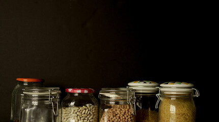 Legumes, lentils, chickpeas and beans assorted in different jars on a black background. Front view.