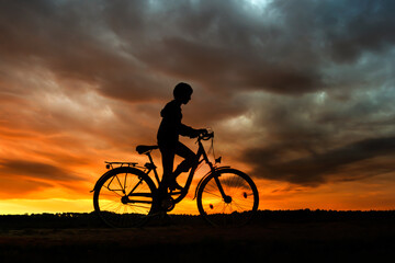 Fototapeta na wymiar Boy , kid 10 years old, riding bike in countryside in dramatic cloudy sky background, silhouette of riding person at sunset in nature