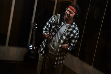 Portrait of young man, male artist looking focused, singing into a condenser microphone while recording a song in a professional studio