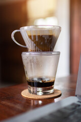 Drip brewing; clear glass of coffee mug and paper dripping bag with ground coffee in soft sunlight at home. Work from home. 