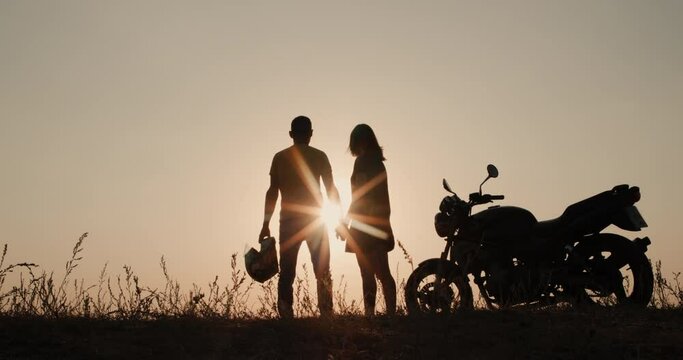 Silhouettes of a young couple near a motorcycle. Standing next to each other at sunset