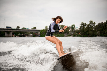 active brown haired woman stands on the wake surf board and rides the wave.