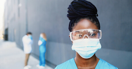 Portrait of African American beautiful woman doctor in medical mask and goggles looking at camera. Close up female physician in respiratory protection. Doctors on background. Zooming in. Dolly shot