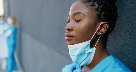 Tired young African American woman doctor taking off medical mask and sipping hot drink while...