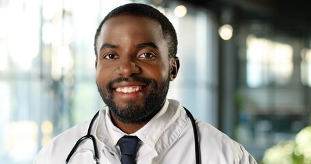 Portrait of African American young man doctor with stethoscope smiling at camera cheerfully. Handsome happy male physician smile. Medic in white gown in clinic. Indoor.