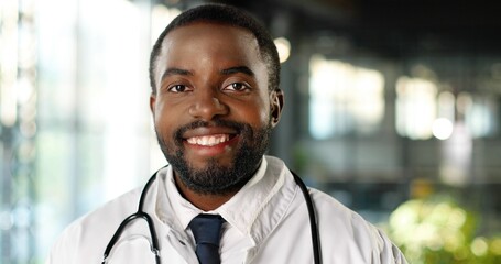 Portrait of African American young man doctor with stethoscope smiling at camera cheerfully. Handsome happy male physician smile. Medic in white gown in clinic. Indoor.