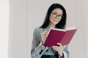 Fototapeta premium Beautiful business teacher plans her lessons, educates high school student, makes notes in notepad, wears spectacles and formal clothes, poses against white background. Woman writes down plans