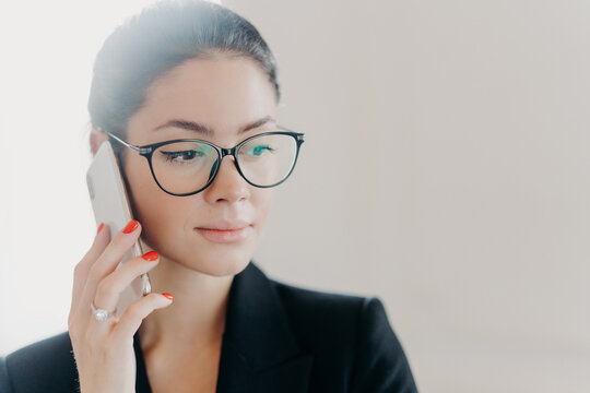 Headshot Of Serious Brunette Female Manager With Makeup Wears Trendy Eyeglasses, Formal Clothes, Has Red Manicure, Calls Business Partner During Remote Job, Poses Indoor Against White Background