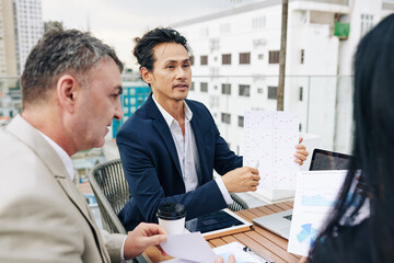 Mixed-race businessman pointing at chart in his hands when talking to his colleagues at meeting
