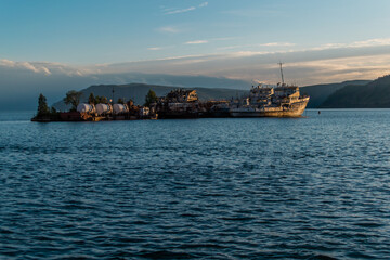 Old abandoned white ships on small island with trees in blue sea in the light of the setting sun against the backdrop of mountains and clouds. Lake Baikal