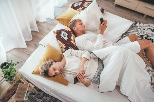 Top view of the couple lying in cozy bed in bedroom and browsing internet and checking mails using each other modern smartphone. Modern technology, social issues and relations problems concept image.
