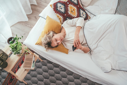 Young blonde female dressed pijama lying in the cozy bed under the blanket and browsing a smartphone internet. Sweet cozy home and wireless technology concept image.