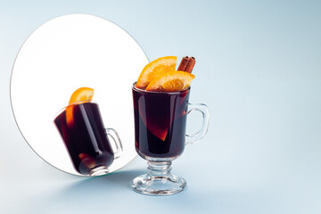 Glass of red glogg or mulled wine with orange slices and cinnamon stick near mirror, on blue background, horizontal, copy space