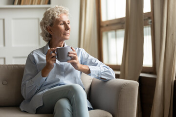 Time for good memories. Pleasant lonely older woman resting on sofa in living room drinking tea or coffee, pensive looking aside remembering life, thinking of current events, enjoying serenity at home
