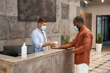 Young male receptionest wearing medical mask standing at reception in the modern office and...