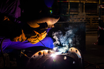 Welder is assembling the workpiece by process shielded metal arc welding (SMAW). Welder in blue uniform, safety shoes, leather gloves, welding mask. He is sitting and welding. Welder, Light and smoke.