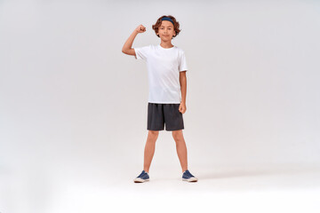 Fototapeta na wymiar Strong kid. Full length of a strong teenage boy in sportswear looking at camera and showing his biceps while posing isolated over grey background in studio