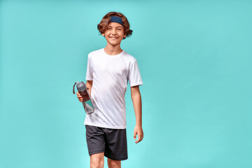 A cute boy with bottle of water looking at camera and smiling, resting after training or workout while standing isolated over blue background