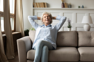 What a good day. Peaceful happy grey-haired old woman relaxing on comfy couch in living room at home, sitting calm with closed eyes smiling and daydreaming, napping, taking break in domestic work