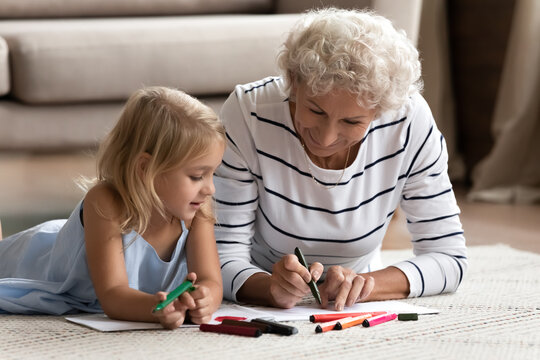 That is how to paint it. Caring mature granny teaching interested granddaughter preschooler to draw sketches on paper sheets, elderly nanny and little girl lying on warm floor indoor coloring pictures
