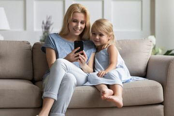 Here we are. Caring grownup elder sister explaining younger girl preschooler how to use mobile webcam app, happy young foster mother showing little adopted daughter funny cartoon online on smartphone
