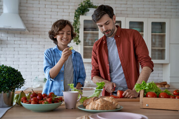 Enjoying weekend at home. Young dad and little happy son cutting fresh vegetables, preparing a salad together while standing in the modern kitchen