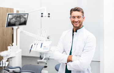 Confident male dentist standing in his modern clinic. Dental hygienist wearing protective glasses smiling at the camera