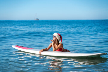A woman in a red bikini and a Santa hat lies on a SUP board with the sea and a yacht floating on the horizon. He looks away and smiles.