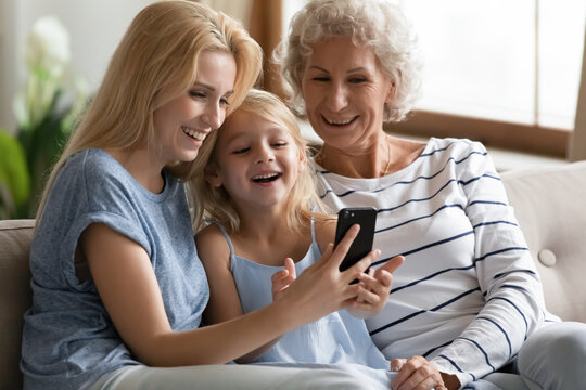 Cute thing. Happy laughing little kid girl relaxing on couch on weekend with senior grandmother and adult mom reading internet blog on cell, playing online game, sharing photos, enjoying video by wifi