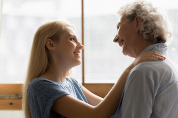 Best mommy in world. Affectionate young millennial daughter hugging silver-haired older mother with tender and care, happy adult grandkid gently holding hands on shoulders of beloved granny, side view