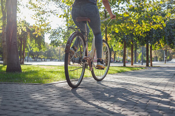 A girl in jeans and sneakers rides a beige bike in the park among green trees and lawn