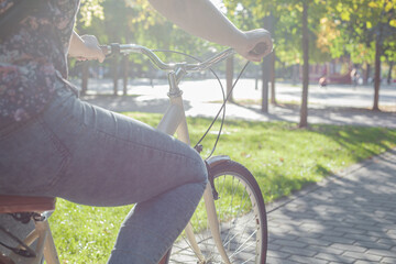 Fototapeta na wymiar A girl in jeans and sneakers rides a beige bike in the park among green trees and lawn