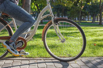 Fototapeta premium A girl in jeans and sneakers rides a beige bike in the park among green trees and lawn