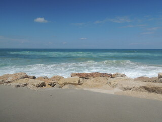 Seashore, turquoise sea, white waves, blue sky, sand and stones.