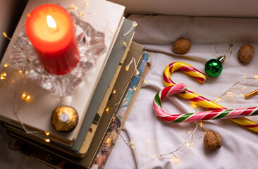 Christmas or New Year's comfort. Composition with a stack of books, a red candle, seasonal sweets, Christmas tree decorations and lights. Home atmosphere, relaxation, winter holidays