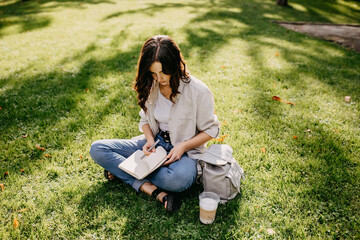 Young woman student sitting on grass in a park with notebook, taking notes.