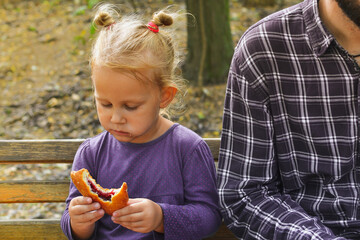 Baby and dad are sitting on a bench in an autumn park. The child eats a bun outdoors.