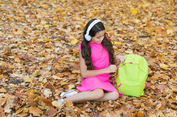 girl in earphones with school bag. kid listen music in autumn park. fall is a time for study. back to school. concept of online education. child sit in autumn leaves