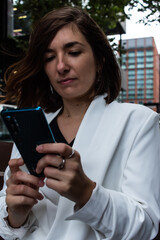 Fototapeta na wymiar Attractive young female wearing a white suit and using her phone while sitting on a bench and waiting to start a meeting at her office in London, United Kingdom