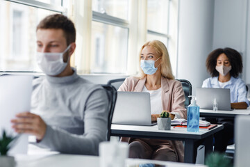 Teamwork on project. Multiracial employees in protective masks work on laptops at desks with antiseptics, focus on lady, looking out the window