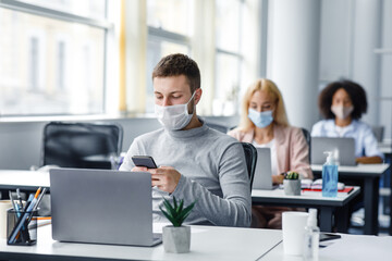 Fototapeta na wymiar Modern technology for work during quarantine covid-19. Young man manager in protective mask typing on smartphone at workplace with laptop