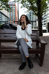 Fototapeta premium Attractive young female wearing a white suit and talking on the phone while sitting on a bench waiting to start a meeting at her office in London, United Kingdom