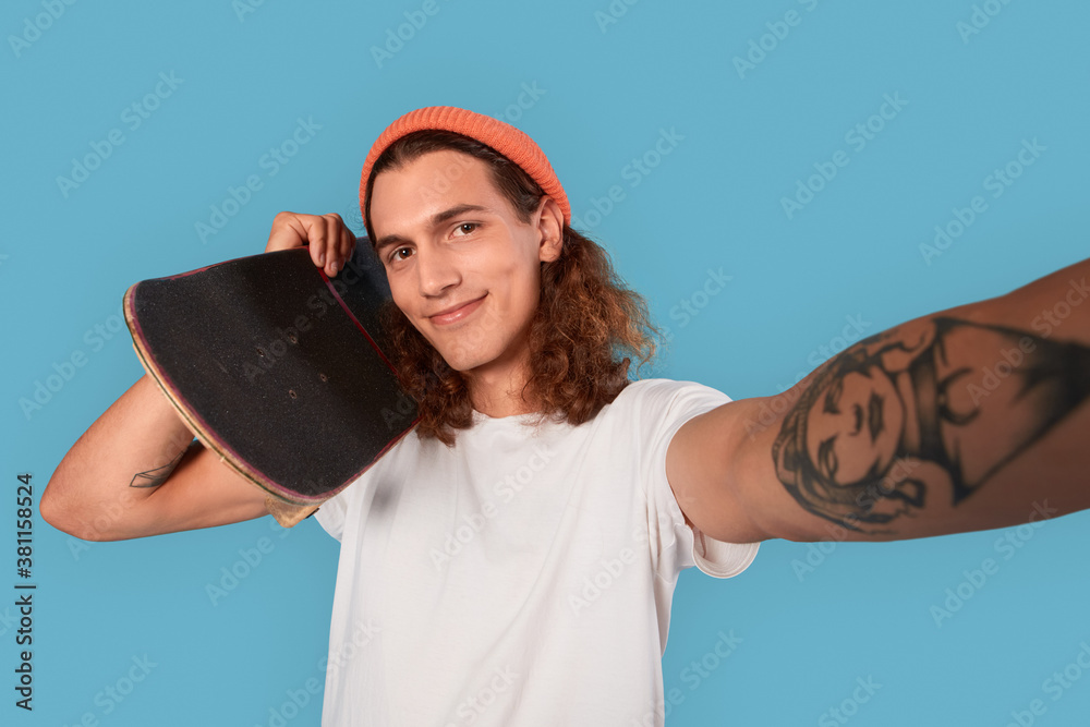 Wall mural Young adult guy holding skateboard, making selfie